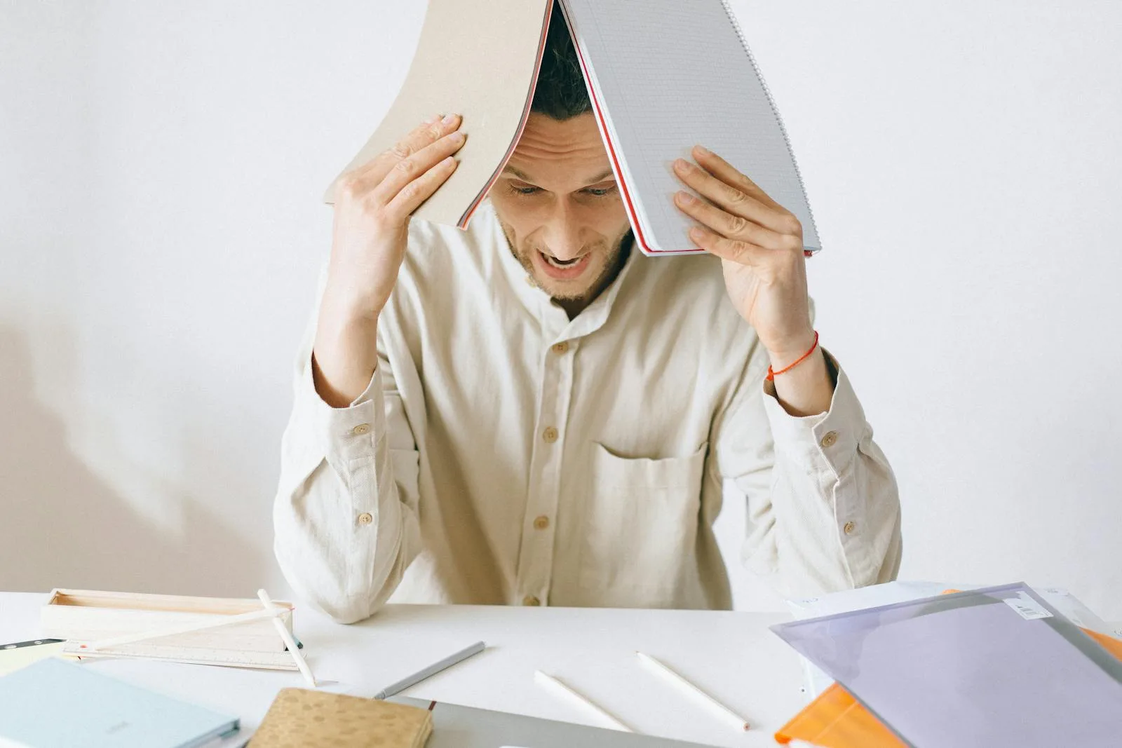 A stressed adult male worker sits at a desk with open notebooks, exhibiting signs of frustration and burnout.
