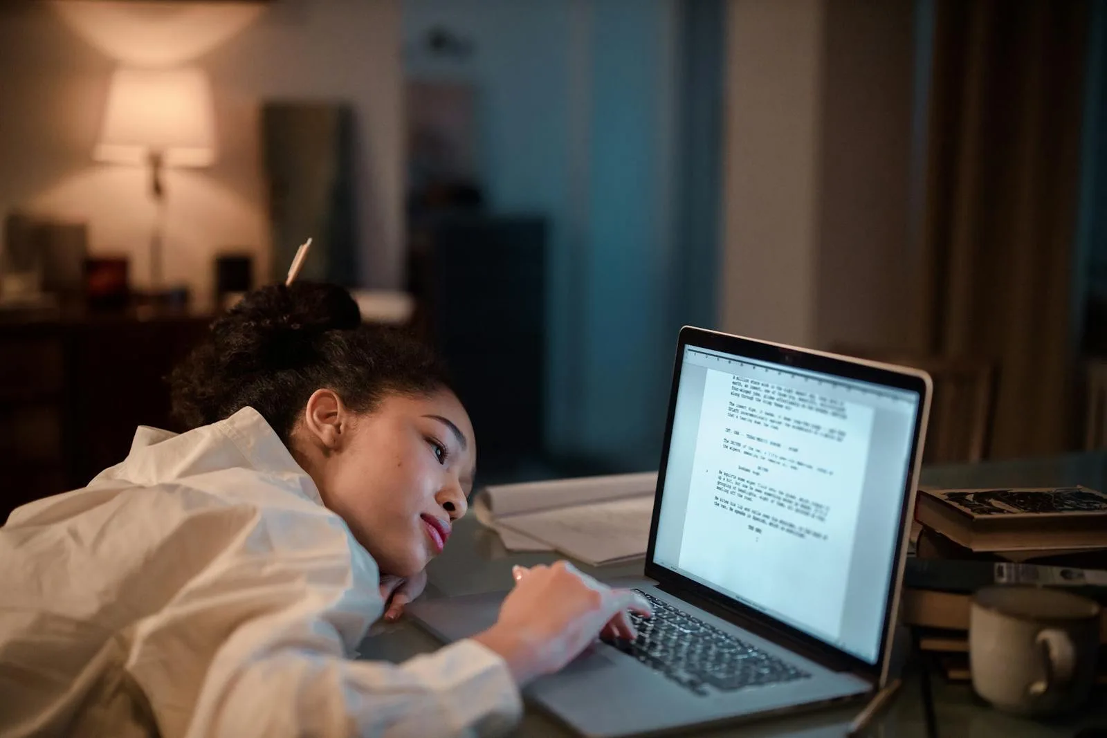 A woman in casual clothing lying on a desk, working late on a laptop at home.
